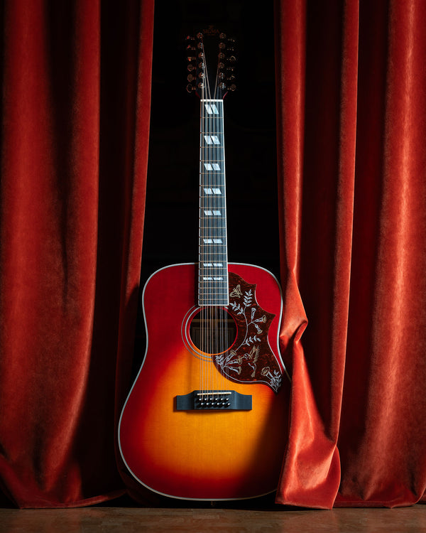 Sigma Guitar with a Gibson Hummingbird Style Sunburst and Pickguard, peeking out from red velvet curtains